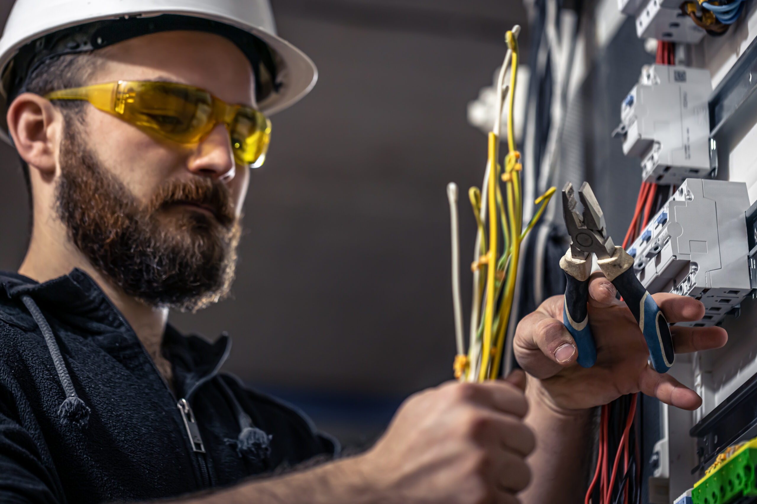 A male electrician works in a switchboard with an electrical connecting cable, connects the equipment with tools.
